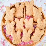 A round wooden tray holds bunny-shaped vegan cookies surrounded by colorful sprinkles, with hints of banana and peanut butter delighting the senses.