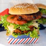 Vegan black bean burger with a sesame bun, topped with fresh lettuce, tomato, cheese, and sauce, all served in a stylish striped container.