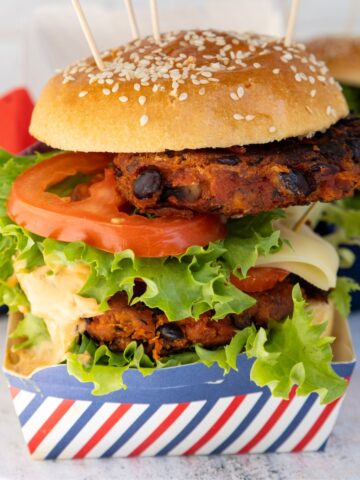 Vegan black bean burger with a sesame bun, topped with fresh lettuce, tomato, cheese, and sauce, all served in a stylish striped container.