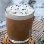 A clear glass mug filled with a frothy latte topped with whipped cream and sprinkled with dried lavender buds. The mug is on a woven coaster.
