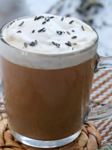 A clear glass mug filled with a frothy latte topped with whipped cream and sprinkled with dried lavender buds. The mug is on a woven coaster.