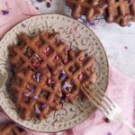 A plate of dark chocolate waffles topped with pink and purple flower petals sits on a pink napkin, with a fork resting on the plate.