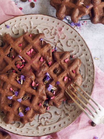 A plate of dark chocolate waffles topped with pink and purple flower petals sits on a pink napkin, with a fork resting on the plate.