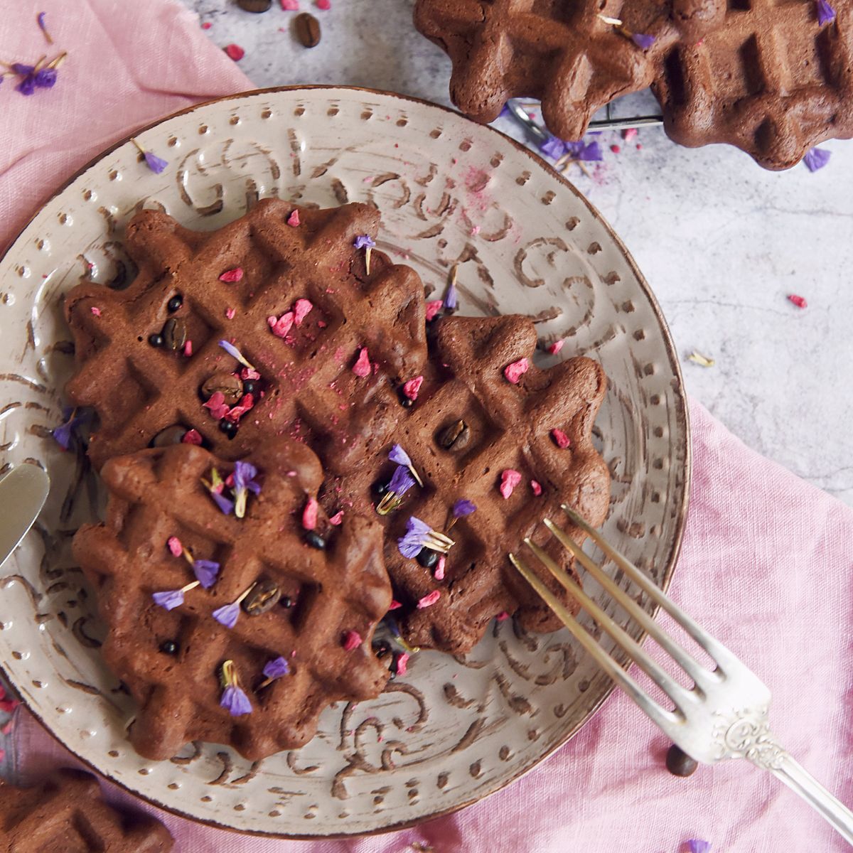 A plate of dark chocolate waffles topped with pink and purple flower petals sits on a pink napkin, with a fork resting on the plate.