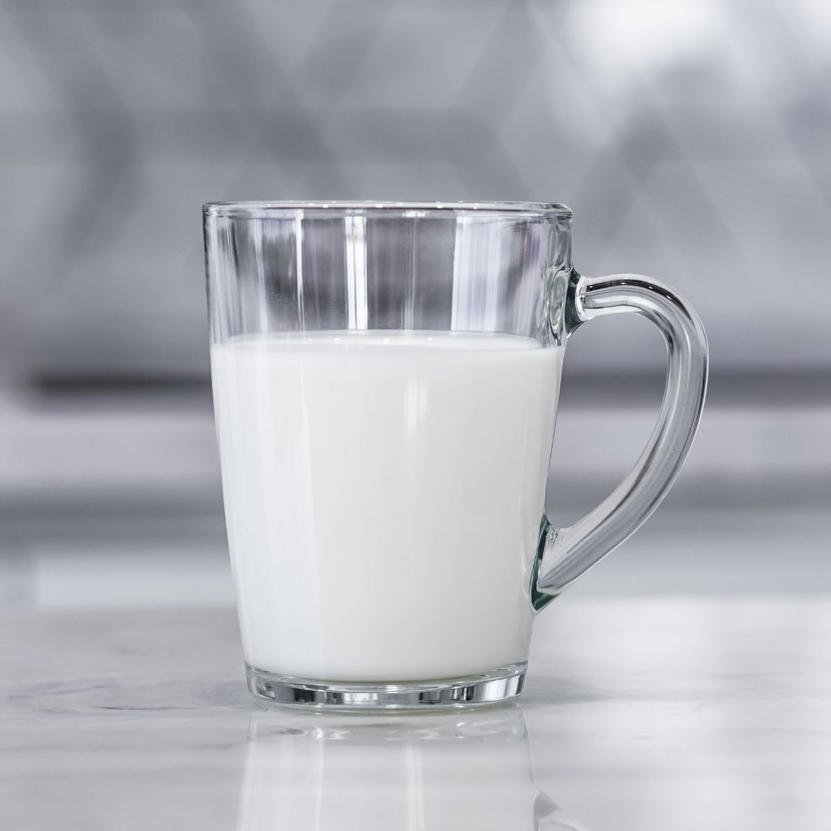 A clear glass mug filled with milk sits on a marble countertop, beside a plate of dark chocolate protein waffles, against a blurred gray geometric patterned background.