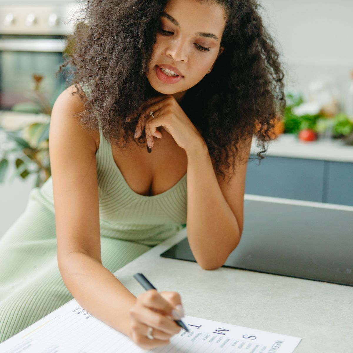 Woman in a green dress writing on a piece of paper in a kitchen, crafting her Free Easy 30 Day Vegan Meal Plan For Beginners