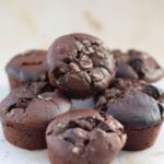 A close-up of six chocolate protein muffins stacked on a light surface.