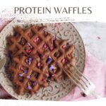 A plate with vegan protein waffles is drizzled with rich dark chocolate and garnished with delicate edible flowers, all set on a pink napkin. Text reads: "Easy Vegan Dark Chocolate Protein Waffles" and "wemadethisvegan.com".