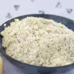 A blue bowl filled with a crumbly mixture, likely vegan crumble topping, sits next to two apples on a white surface with a patterned background.