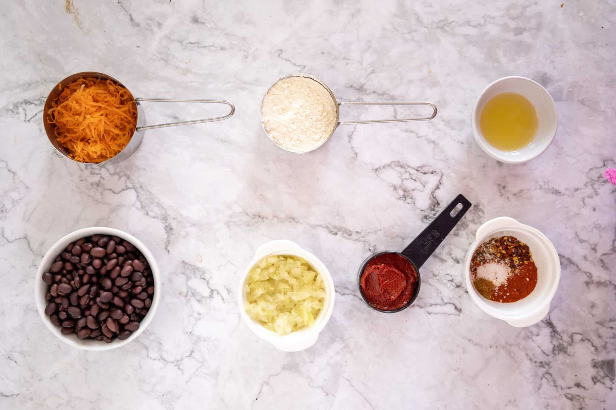 Vegan Black Bean and Carrot Burger Recipe 161 Ingredients neatly arranged on a marble surface in individual bowls: black beans, shredded cheese, chopped onions, tomato paste, flour, mixed spices, and a cup of oil.