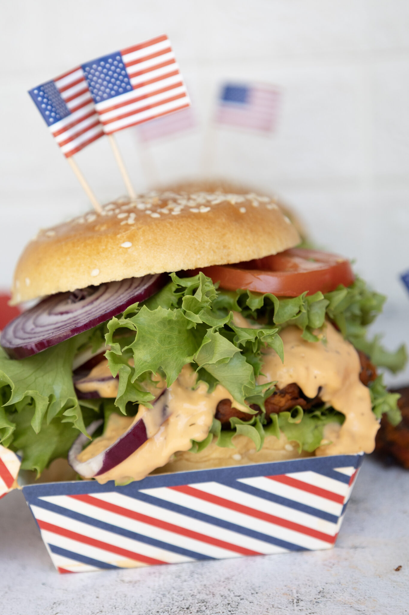 A burger with lettuce, tomato, red onion, and sauce in a bun, topped with small American flags, in a striped paper container.