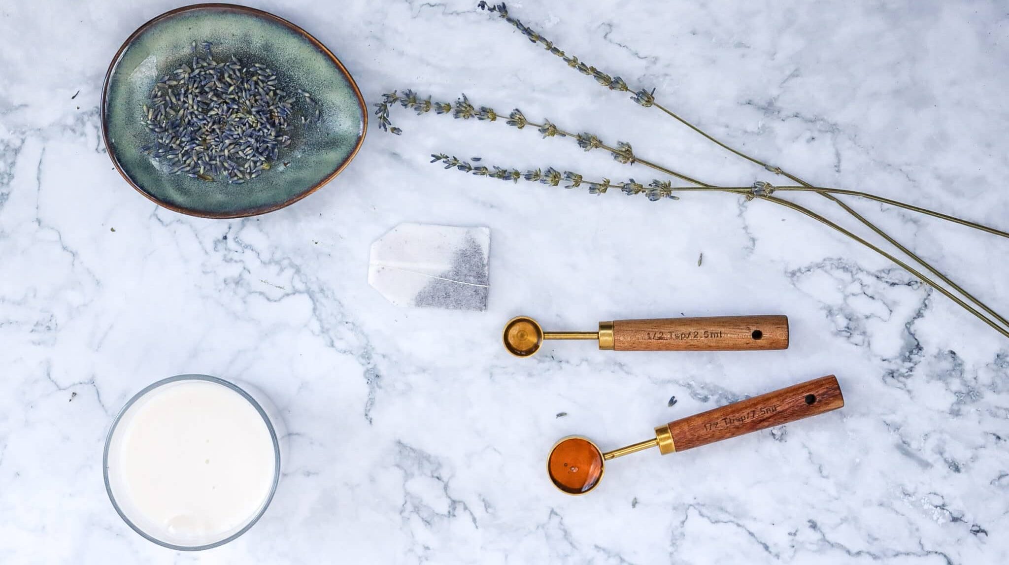 Simple Vegan Lavender London Fog Latte Recipe 121 A marble surface with lavender sprigs, a bowl of lavender buds, a tea bag, a candle, and two wooden scoops with powder.