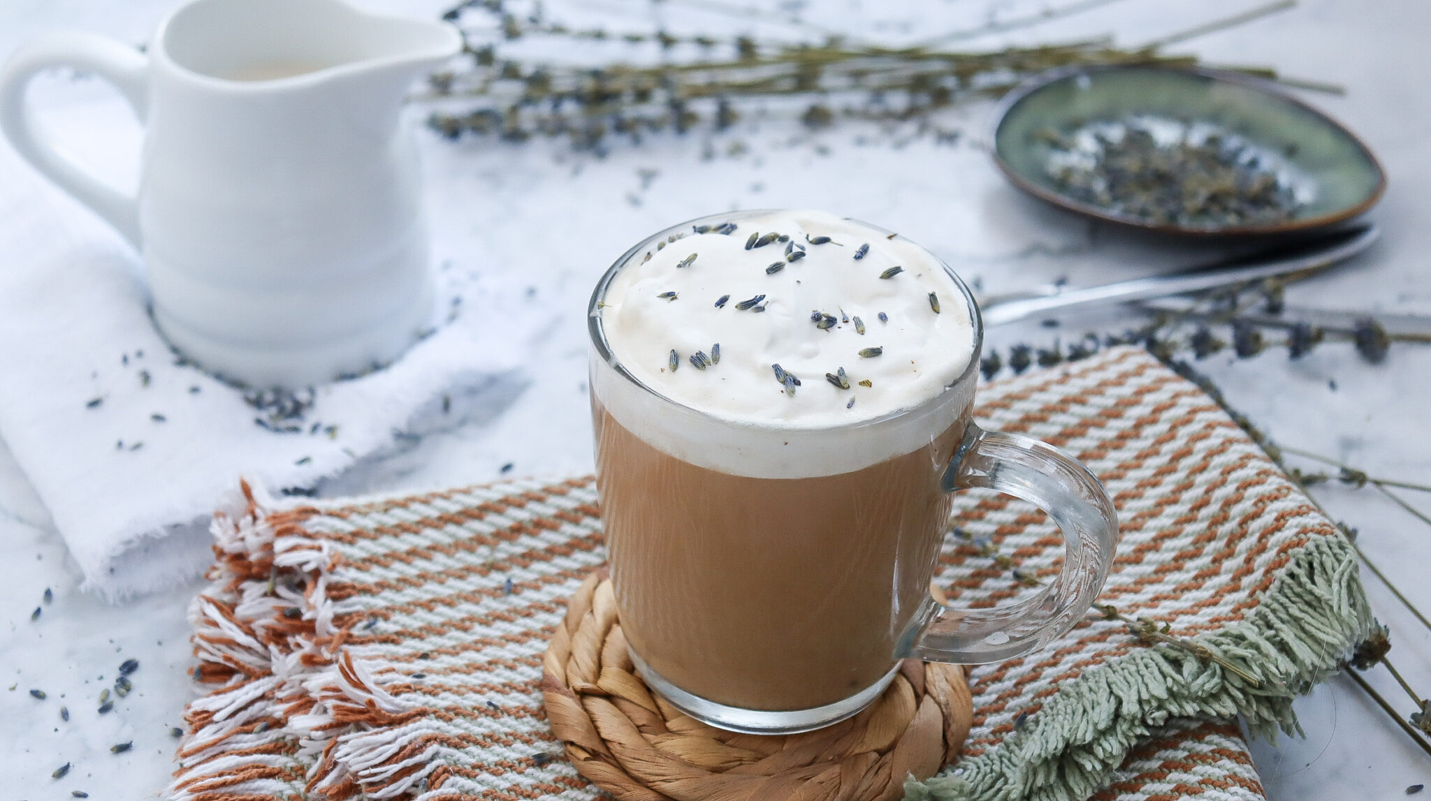 Glass mug of creamy coffee topped with lavender sprinkles on a woven coaster, placed on a striped cloth. A small pitcher and a green saucer with lavender are in the background.