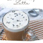 A vegan lavender London fog latte, crowned with frothy cream and a sprinkle of lavender buds, sits elegantly in a clear mug on a woven mat. Explore more at wemadethisvegan.com.