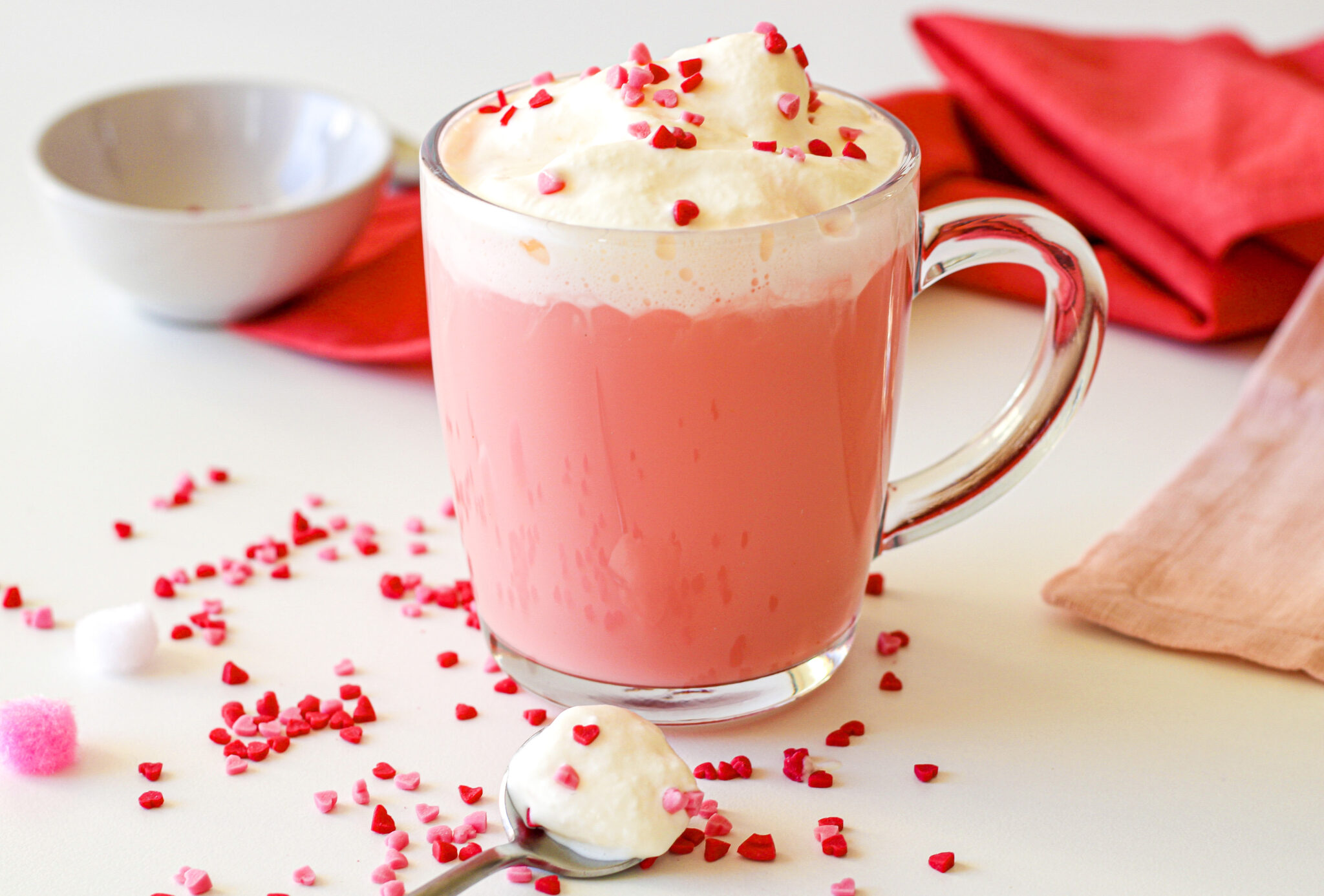 A mug of pink hot chocolate topped with whipped cream and heart-shaped sprinkles. A spoon with whipped cream is in the foreground, and red napkins are in the background.