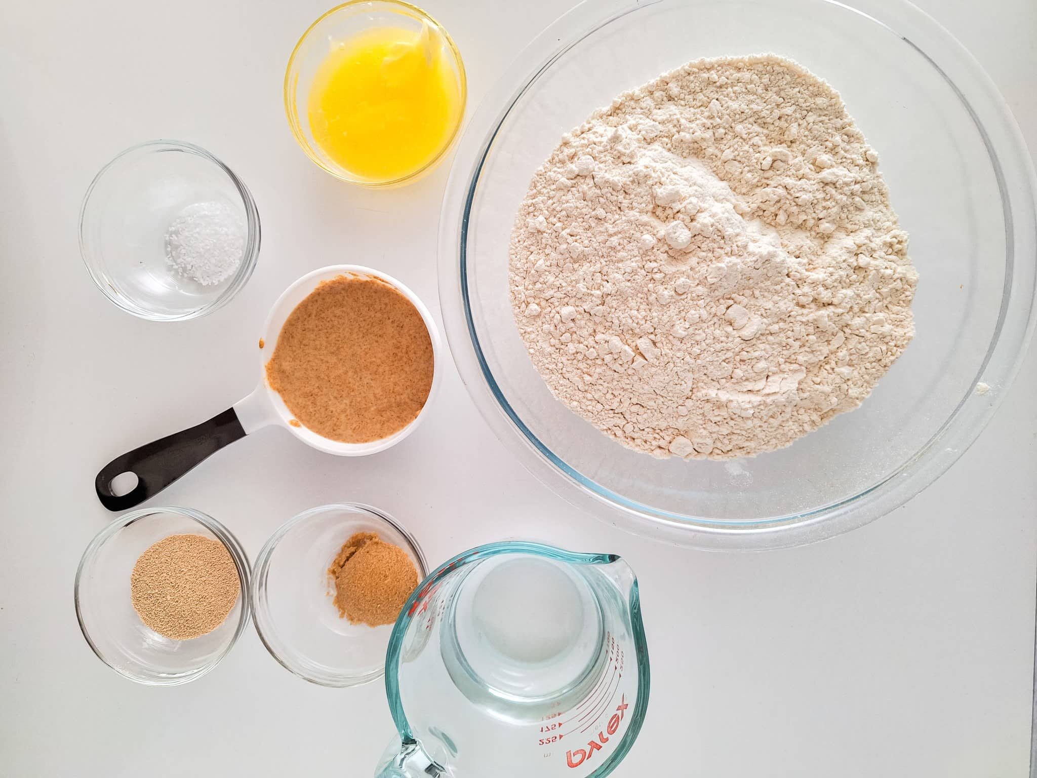 Sourdough Discard Pretzels Recipe 351 Ingredients for baking: flour in a bowl, melted butter in a glass, small bowls of sugar, yeast, and salt, and a measuring cup with water, all arranged on a white surface.