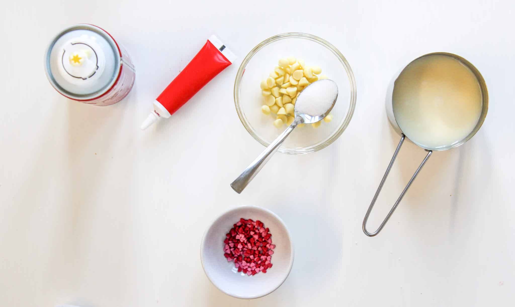 Red Velvet Hot Chocolate Recipe 125 Baking ingredients on a white surface: milk in a measuring cup, white chocolate chips with sugar in a bowl, red icing tube, pink sprinkles in a bowl, and a whipped cream can.