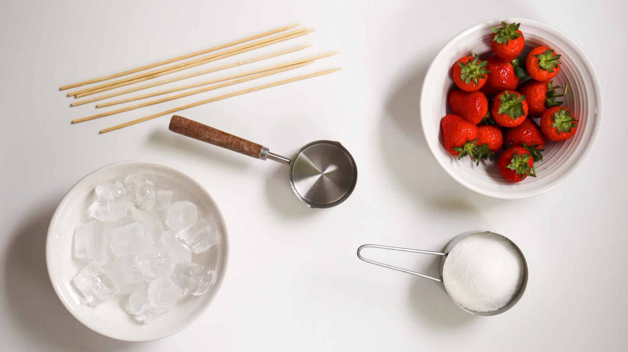 Strawberry Tanghulu Recipe (How To Make Candied Strawberries) 173 Bowls of strawberries, sugar, and ice cubes are arranged on a table with wooden skewers and an ice cream scoop.