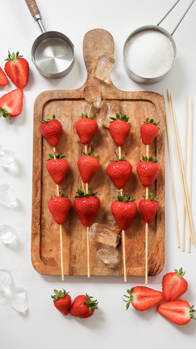 Fresh strawberries threaded onto wooden skewers on a wooden board, surrounded by halved strawberries, ice cubes, skewers, and measuring cups with sugar.