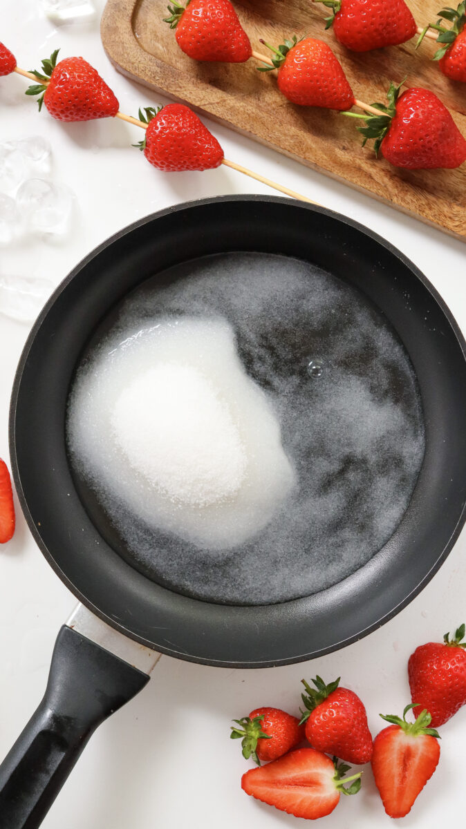 A frying pan with granulated sugar melting inside, surrounded by strawberries, both whole and halved, and pieces of ice on a white surface.