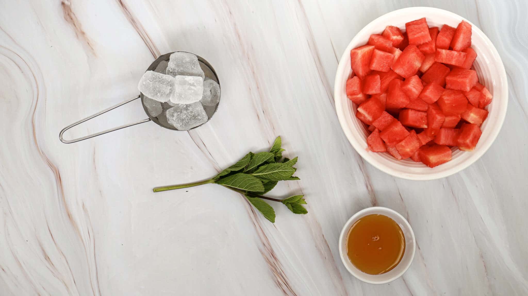 Watermelon Slushie Recipe 102 Bowl of cubed watermelon, a small bowl of honey, ice cubes in a measuring cup, and a sprig of mint on a marble surface.