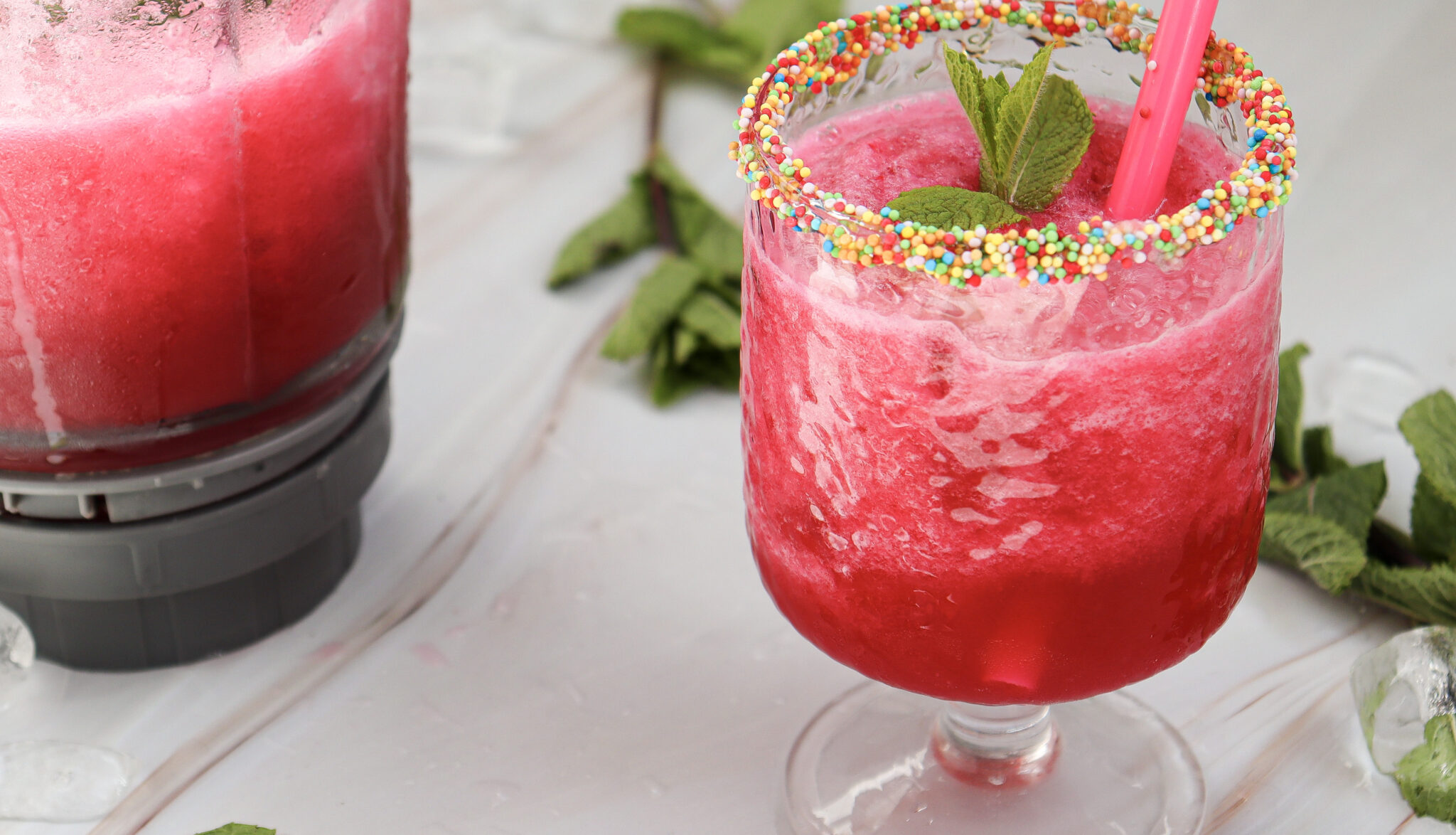 A glass of pink slush with a colorful sprinkle rim, garnished with mint leaves and a straw, next to a blender.