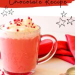 A glass mug of red velvet hot chocolate topped with whipped cream and red sprinkles on a white surface with a red napkin. Text reads: "Red Velvet Hot Chocolate Recipe.
