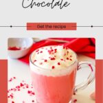 Glass mug of red velvet hot chocolate topped with whipped cream and red sprinkles, set on a white surface with a red cloth in the background.
