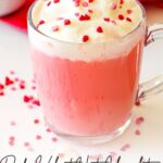 Clear glass mug filled with pink hot chocolate topped with whipped cream and heart sprinkles, placed on a white surface with red accents. Text overlay reads, "Red Velvet Hot Chocolate Recipe.