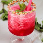 A refreshing watermelon slushie features a red slushy drink in a glass with a candy sprinkle rim, garnished with mint leaves and finished with a pink straw.