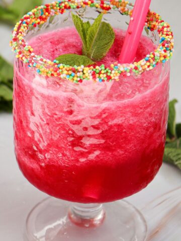 A refreshing watermelon slushie features a red slushy drink in a glass with a candy sprinkle rim, garnished with mint leaves and finished with a pink straw.