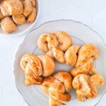 A plate of seven golden-brown sourdough discard pretzels sits on a table, next to a small bowl of sugar-coated chunks.