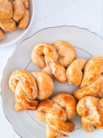 A plate of seven golden-brown sourdough discard pretzels sits on a table, next to a small bowl of sugar-coated chunks.