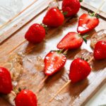 Strawberry Tanghulu Recipe - Candied strawberries on skewers rest on a wooden tray, complemented by melting ice cubes.
