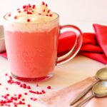 A glass mug filled with pink hot chocolate, topped with whipped cream and heart-shaped sprinkles. Two spoons rest on a pink napkin beside the mug.