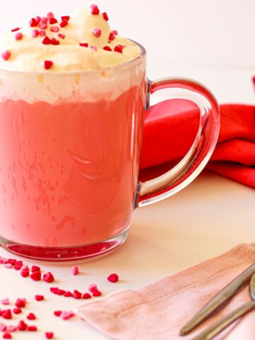 A glass mug filled with pink hot chocolate, topped with whipped cream and heart-shaped sprinkles. Two spoons rest on a pink napkin beside the mug.