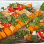 Sliced roasted butternut squash drizzled with ginger peanut sauce, adorned with sliced red chili, shallots, and fresh cilantro in a pristine white baking dish.
