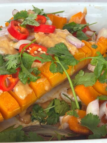Sliced roasted butternut squash drizzled with ginger peanut sauce, adorned with sliced red chili, shallots, and fresh cilantro in a pristine white baking dish.