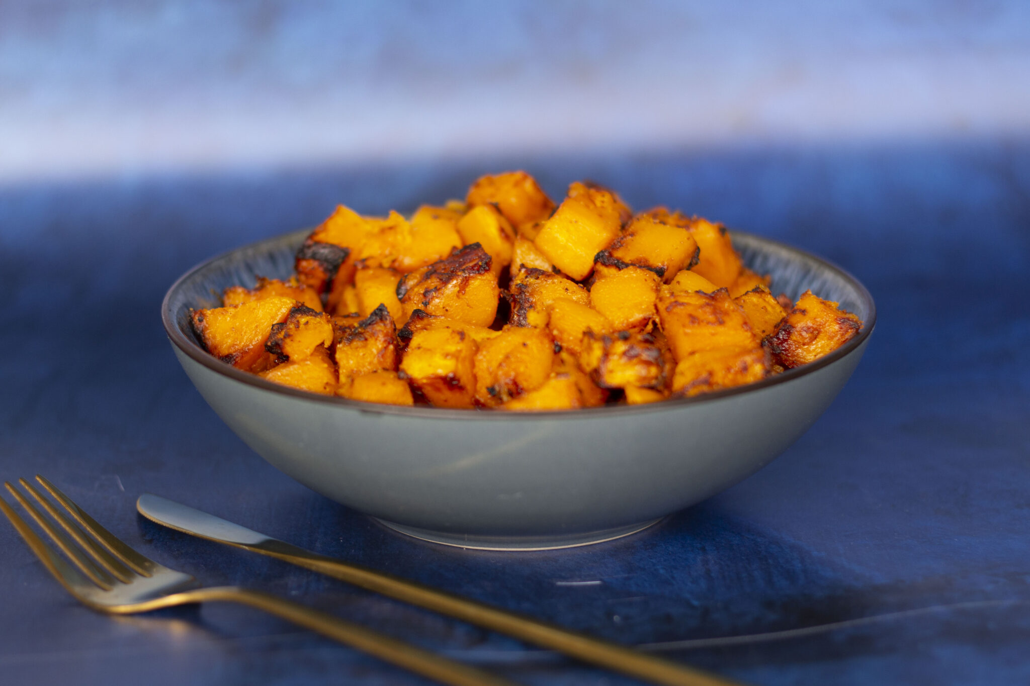 A bowl filled with roasted cubes of butternut squash against a blue background, with a gold fork and knife placed beside it.