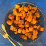 A bowl of roasted cubed butternut squash seasoned with herbs and spices, accompanied by a gold fork and knife on a blue surface.