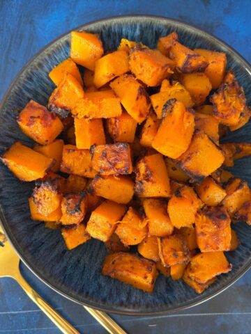 A bowl of roasted cubed butternut squash seasoned with herbs and spices, accompanied by a gold fork and knife on a blue surface.