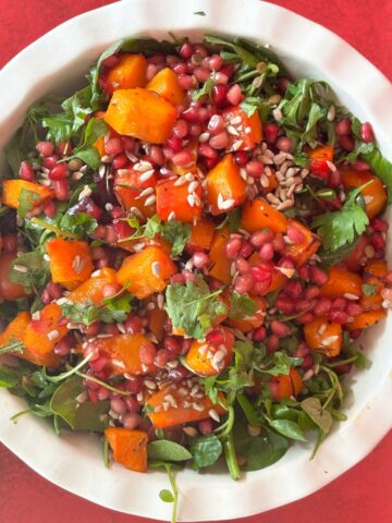 A delightful beetroot and butternut squash salad featuring arugula, pomegranate seeds, cilantro, and sunflower seeds is elegantly presented in a round bowl on a vibrant red surface.