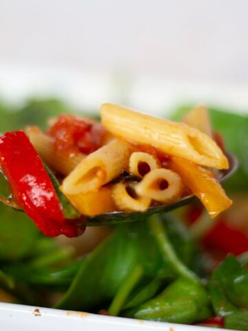 Close-up of a spoonful of Butter Bean Pasta salad with spinach, roasted red and yellow peppers, and penne pasta over a background of additional salad.