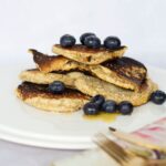 A stack of oat flour pancakes topped with blueberries sits on a white plate, with syrup drizzled on top. Utensils are in the foreground.