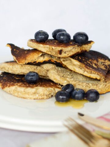 A stack of oat flour pancakes topped with blueberries sits on a white plate, with syrup drizzled on top. Utensils are in the foreground.