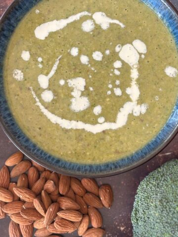 A bowl of creamy broccoli soup with a swirl of cream, surrounded by almonds, sits enticingly on a rustic surface.