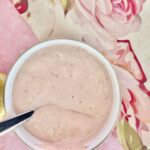 A bowl of creamy light pink vegan yoghurt sauce with a spoon inside, placed on a floral-patterned tablecloth.