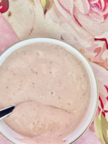 A bowl of creamy light pink vegan yoghurt sauce with a spoon inside, placed on a floral-patterned tablecloth.
