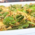 Close-up of creamy vegan pasta with spinach leaves, sun-dried tomatoes, and red pepper flakes in a white dish.
