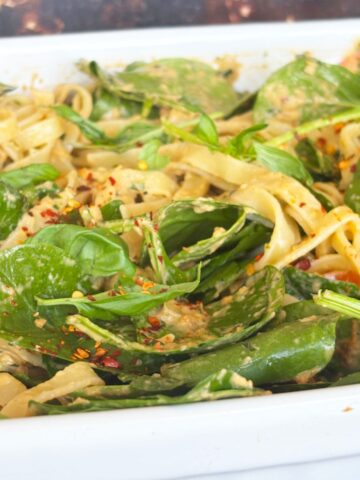Close-up of creamy vegan pasta with spinach leaves, sun-dried tomatoes, and red pepper flakes in a white dish.